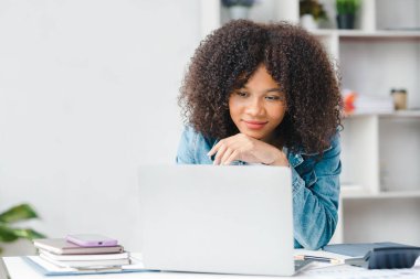 American teenage woman sitting in white office with laptop, she is a student studying online with laptop at home, university student studying online, online web education concept.
