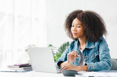 American teenage woman sitting in white office with laptop, she is a student studying online with laptop at home, university student studying online, online web education concept.