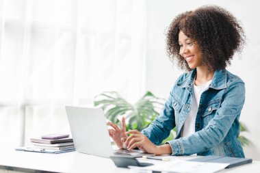 American teenage woman sitting in white office with laptop, she is a student studying online with laptop at home, university student studying online, online web education concept.