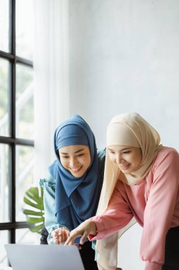 Two hijab-clad Asian women are brainstorming together in a conference room for a startup, run by a young, talented woman. The management concept runs the company of female leaders to grow the company.