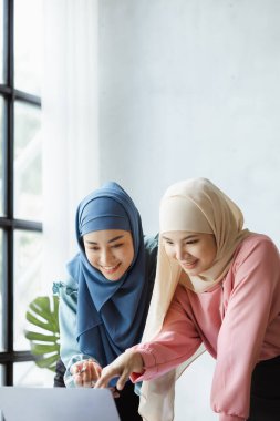 Two hijab-clad Asian women are brainstorming together in a conference room for a startup, run by a young, talented woman. The management concept runs the company of female leaders to grow the company.