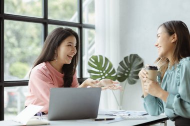 Girişim şirketinin atmosferinde, iki kadın çalışan, satışları artırmak ve yöneticilere raporlar hazırlamak için özetler ve pazarlama planları üzerinde çalışmak üzere fikir alışverişinde bulunuyorlar..