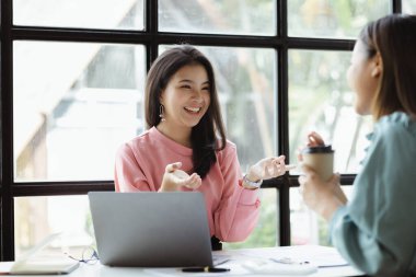 Girişim şirketinin atmosferinde, iki kadın çalışan, satışları artırmak ve yöneticilere raporlar hazırlamak için özetler ve pazarlama planları üzerinde çalışmak üzere fikir alışverişinde bulunuyorlar..