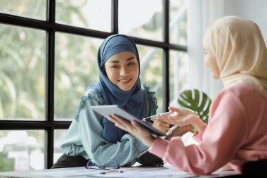 Two hijab-clad Asian women are brainstorming together in a conference room for a startup, run by a young, talented woman. The management concept runs the company of female leaders to grow the company.