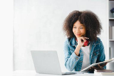 American teenage woman sitting in white office with laptop, she is a student studying online with laptop at home, university student studying online, online web education concept.