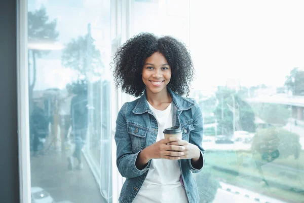 An American woman stands by a window with a cup of hot paper coffee in a startup company meeting room, she is an employee of an international marketing company, she attends a company meeting.