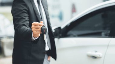 Takım elbiseli bir adam beyaz bir arabanın yanında arabanın anahtarlarını tutuyor ve galeride satıcı olan yeni bir araba alıyor. Araba ticareti kavramı.
