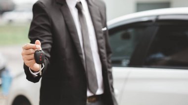 Takım elbiseli bir adam beyaz bir arabanın yanında arabanın anahtarlarını tutuyor ve galeride satıcı olan yeni bir araba alıyor. Araba ticareti kavramı.