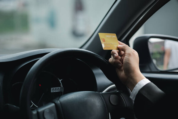 An Asian man in a formal suit is in a car, he holds a credit card, he drives into a gas station and pays his credit card bills, travel by car, safe driving, respecting traffic rules.