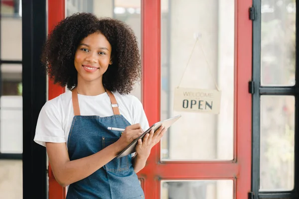 A young American woman stands in front of a restaurant door with an open sign, she is a waitress of a fast food restaurant preparing to open a shop to serve customers. Restaurant concept.