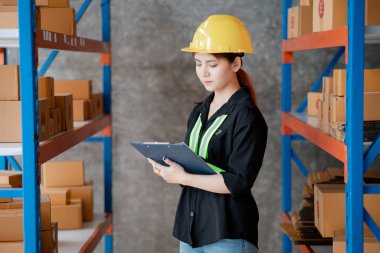 Güvenli kıyafetler giyen Asyalı kadın işçi bir depoda nakliyeden önce depodaki malları kontrol ediyor. Depoda çalışmak ve depoda çalışmak güvenli..