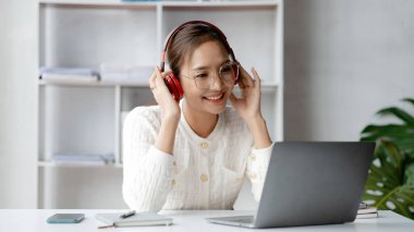 İşten çıkarken müzik dinlemek için kulaklık takan Asyalı kadın, pazarlama departmanında yeni açılan şirketin ofisinde, işe başlamadan önce dinlenmek için zaman ayırıyor..