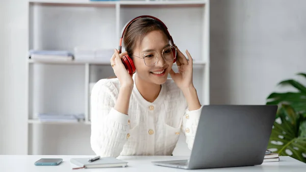 Asian woman wearing headphones to listen to music in her break from work, she is in the office of startup company in the marketing department office, she takes time to rest before starting work again.
