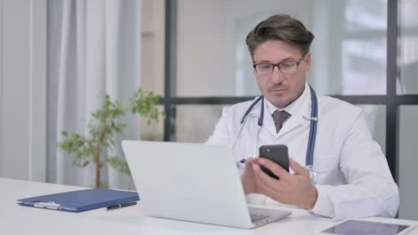 Arts met behulp van Smartphone tijdens het werken op Laptop in de kliniek — Stockvideo