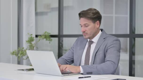 Uomo di mezza età guardando la fotocamera durante l'utilizzo del computer portatile in ufficio — Video Stock