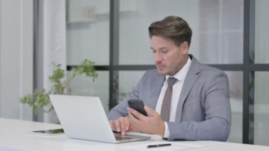 Ofiste dizüstü bilgisayar kullanırken akıllı telefon kullanan orta yaşlı adam 