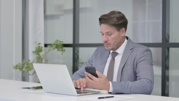 Middelbare leeftijd Man met behulp van Smartphone tijdens het gebruik van Laptop in Office — Stockvideo