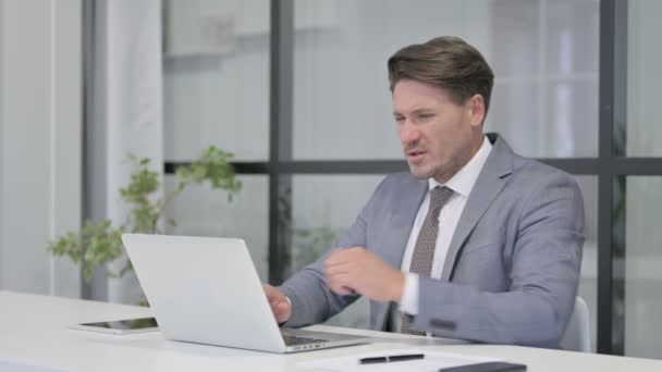 Людина середнього віку, яка відчуває біль у спині, користуючись ноутбуком у офісі — стокове відео