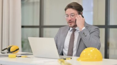 Ofiste dizüstü bilgisayar kullanırken orta yaşlı mühendis telefonla konuşuyor.