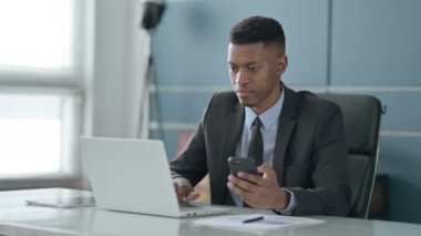 Ofiste dizüstü bilgisayar kullanırken akıllı telefon kullanan Afrikalı iş adamı 