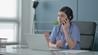 Ofiste dizüstü bilgisayar kullanırken akıllı telefondan konuşan Hintli kadın doktor.