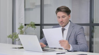 Dizüstü bilgisayar kullanırken Orta Yaşlı Adam Belgeleri Okuyor