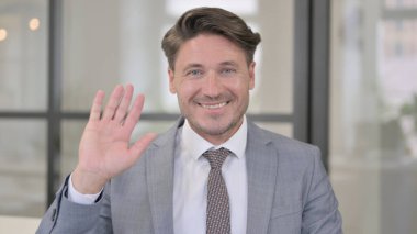 Orta yaşlı adam el sallıyor, hoş geldin diyor.