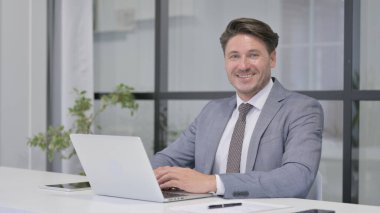 Ofiste dizüstü bilgisayar kullanırken kameraya gülümseyen orta yaşlı adam