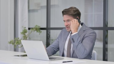 Ofiste dizüstü bilgisayar kullanırken telefonda konuşan kızgın orta yaşlı adam.