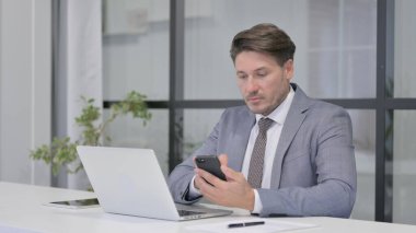 Ofiste dizüstü bilgisayar kullanırken akıllı telefon kullanan orta yaşlı adam