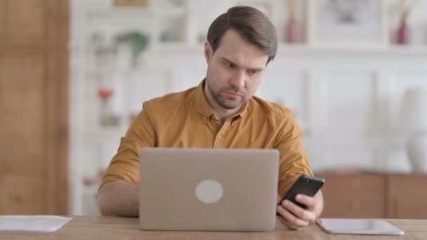 Молодой человек с помощью смартфона во время использования ноутбука в офисе — стоковое видео