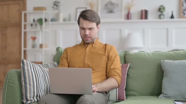 Молодой человек думает во время работы над ноутбуком на диване — стоковое видео