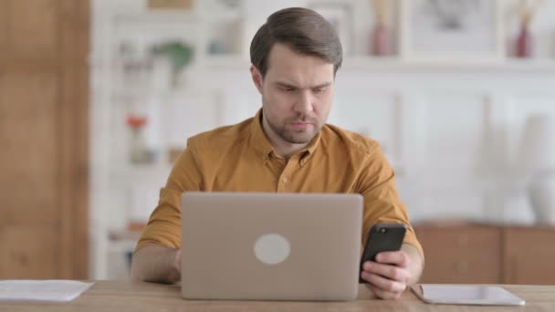 Молодой человек разговаривает по телефону во время использования ноутбука в офисе — стоковое видео