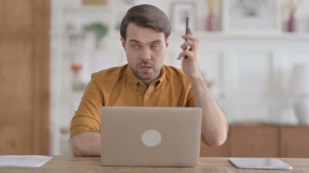 Злой молодой человек разговаривает по телефону во время использования ноутбука в офисе — стоковое видео