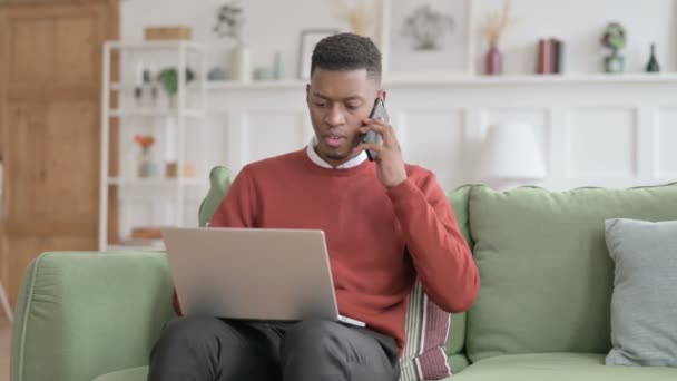 Homme africain en colère avec ordinateur portable parlant sur le téléphone sur le canapé — Video