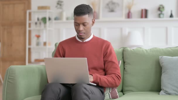 Hombre africano con portátil se siente sorprendido en el sofá — Vídeos de Stock
