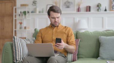 Dizüstü bilgisayarı olan genç adam koltukta akıllı telefon kullanıyor. 