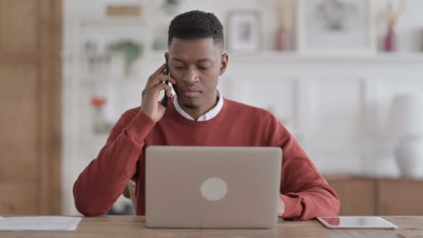 Dühös afrikai férfi beszél telefonon használata közben Laptop az irodában — Stock videók