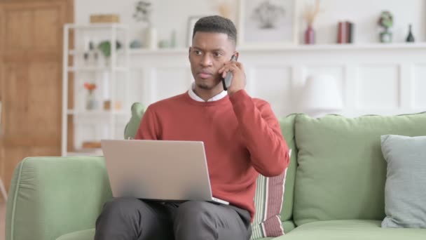 Afrikansk man med laptop talar på telefon på soffan — Stockvideo