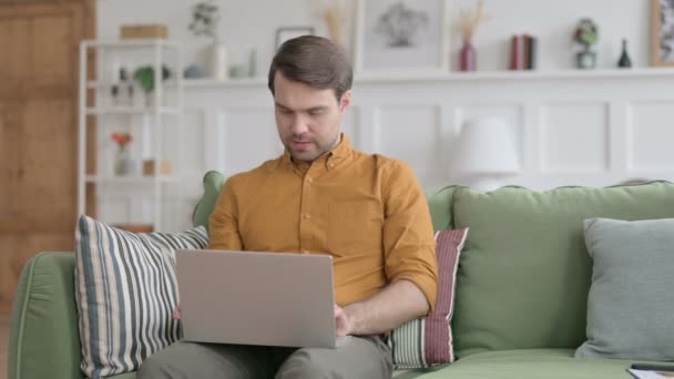 Молодой человек с ноутбуком реагирует на потерю на диване — стоковое видео