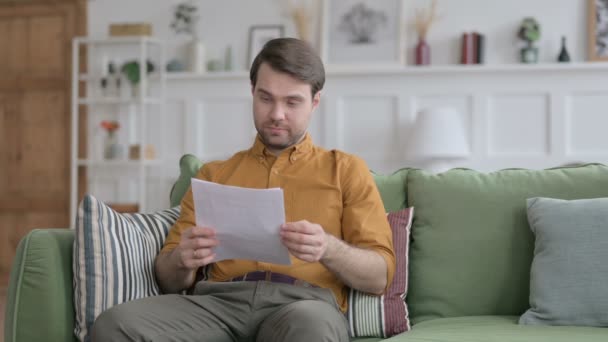 Молодой человек празднует успех на документе на диване — стоковое видео