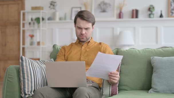 Jonge man met laptop reageert op verlies op documenten, Sofa — Stockvideo