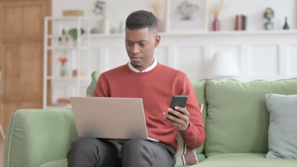 Hombre africano con ordenador portátil usando Smartphone en el sofá — Vídeos de Stock