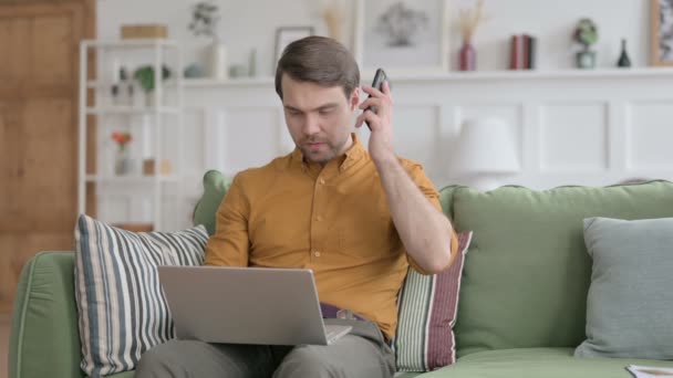 Молодой человек с ноутбуком разговаривает по телефону на диване — стоковое видео