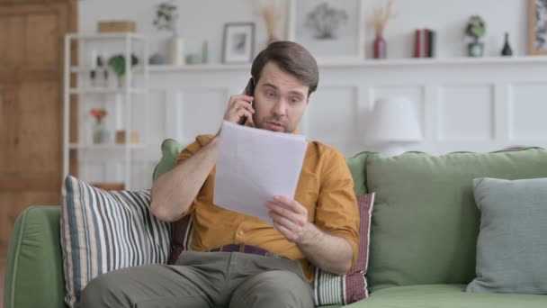 Молодой человек сердится на смартфон во время чтения докладов на диване — стоковое видео