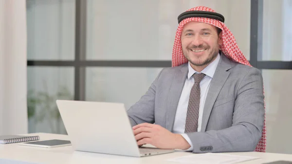 中年阿拉伯商人在办公室使用笔记本电脑时在相机前微笑 — 图库照片