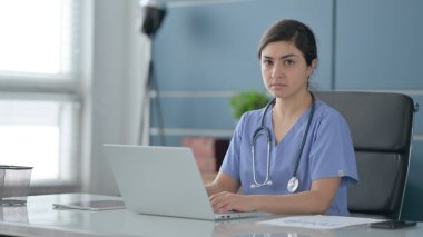 Ofiste dizüstü bilgisayar kullanırken kameraya bakan Hintli kadın doktor