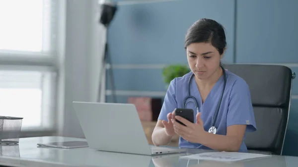 Indiase vrouwelijke arts met behulp van Smartphone tijdens het gebruik van Laptop in Office — Stockfoto