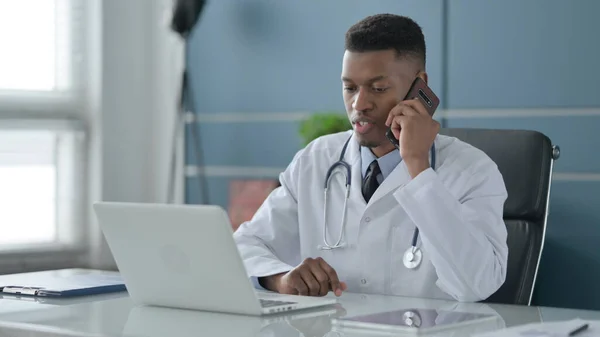 Afrikanska Doctor Talar på smartphone när du använder laptop i Office — Stockfoto