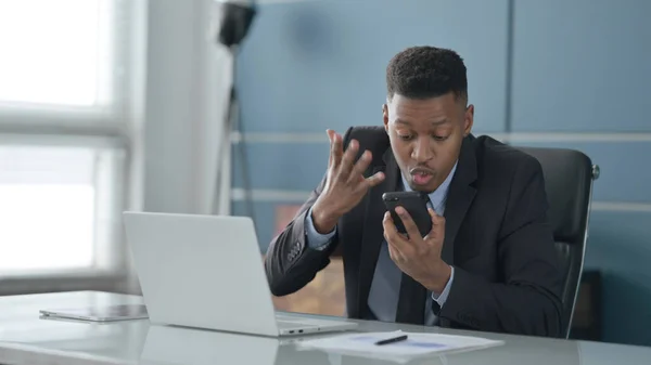 Wütender afrikanischer Geschäftsmann spricht auf Smartphone, während er Laptop im Büro benutzt — Stockfoto